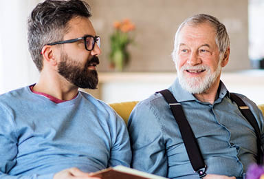 An adult hipster son and senior father with photo album sitting on sofa indoors at home, talking 